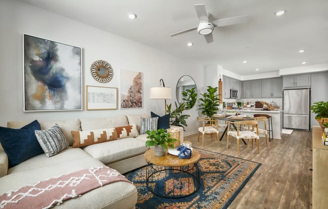 2 BR Apartments in Riverside CA - Lincoln Village - Furnished Living Room with White L-Shaped Couch, Round Wood-Topped Coffee Table, Oriental Rug, White Walls, Ceiling Fan, Dining Table with Chairs, Mounted Circular Mirror, House Plants, and View of Kitchen
