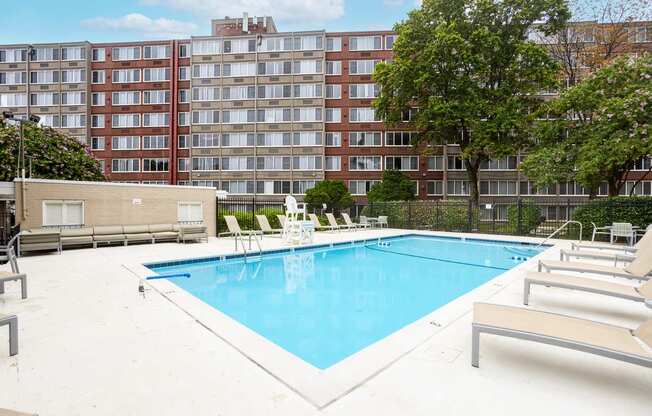 A large swimming pool surrounded by lounge chairs and trees in front of apartment buildings.
