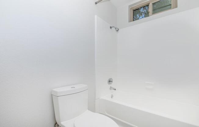 A clean, modern bathroom featuring a white bathtub, showerhead, and a toilet. The walls are painted white, and there is a window above the bathtub allowing natural light to enter the space. The overall design is simple and minimalistic.
