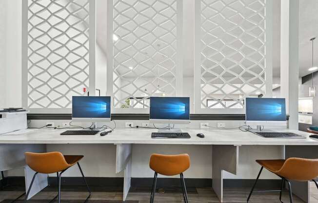 Three computers are on a white desk with orange chairs.