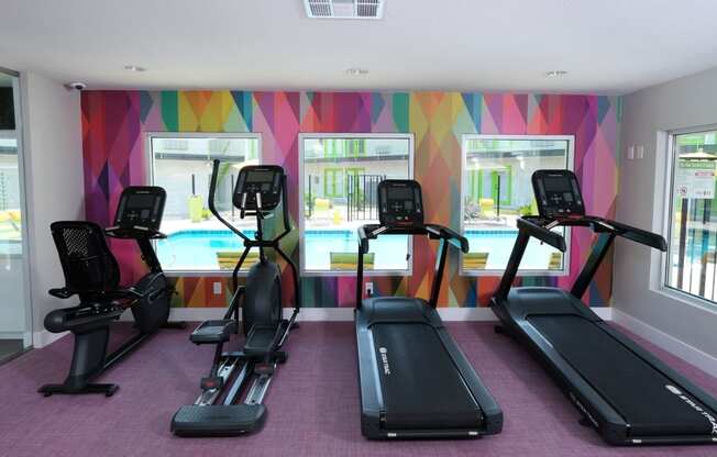 Cardio machines looking out three large windows in a colorful wall in the fitness center at Fusion Las Vegas Apartments.