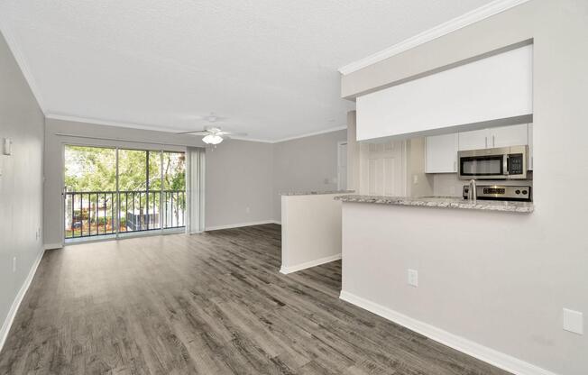Spacious, light-filled living area with modern decor, featuring a ceiling fan and large windows that open to a balcony view. The open-plan kitchen includes a granite countertop and stainless steel appliances, complementing the contemporary style of the room.