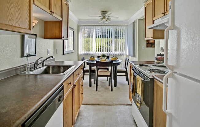 A bright kitchen here at Andorra showcasing warm wood cabinetry, spacious countertops, a double-basin sink, and a full appliance setup, flowing into a cozy dining area with a round table set for four, a ceiling fan overhead, and a large window with vertical blinds that brings in natural light and views of the surrounding green space.