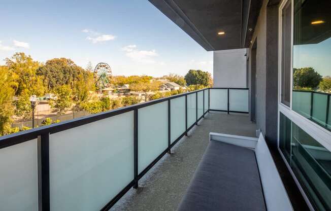 A balcony with a glass railing and a bench overlooks a park with a Ferris wheel.