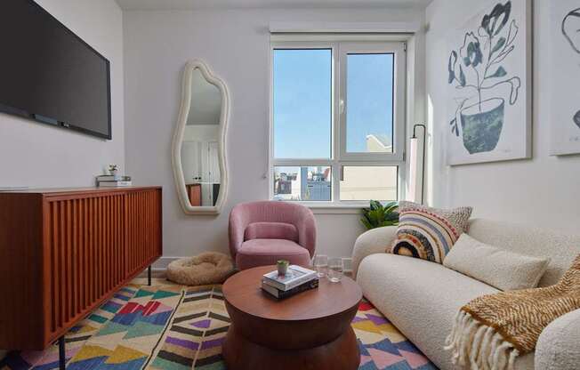 A living room with a white couch, a brown coffee table, a pink armchair, a wooden cabinet, a mirror, a flat-screen TV, and a colorful rug.