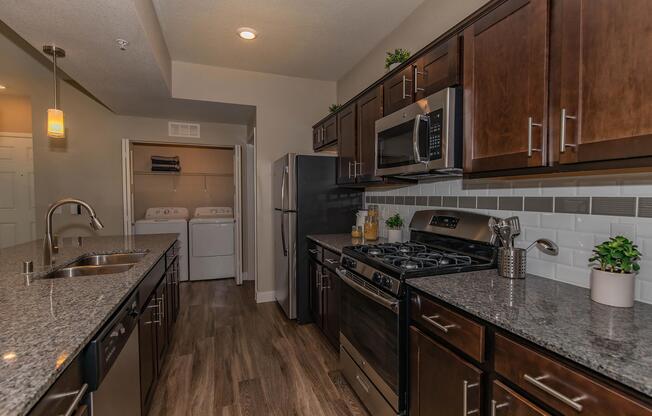 A modern kitchen featuring dark wood cabinets, a stainless steel gas stove, and a microwave. The countertops are made of dark granite, and there is a double sink. In the background, a laundry area is visible with a washer and dryer. The space is well-lit with warm overhead lighting and small decorative plants.