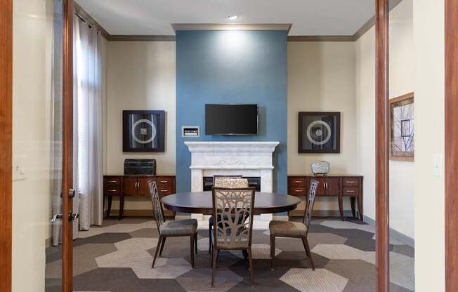 A game lounge here at Landing at Mansfield with a round table and four chairs, blue accent fireplace wall, wall-mounted TV, wood side cabinets, and patterned hexagon carpet.