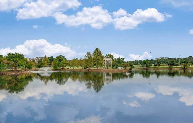 A serene lake with a fountain in the middle, surrounded by trees and a clear blue sky.