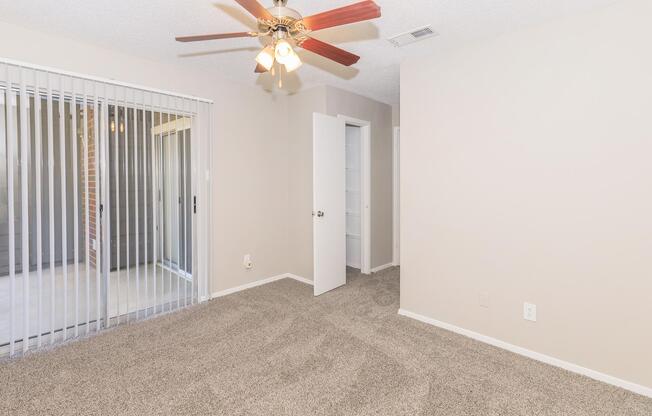 A spacious room with beige walls and plush carpet. A ceiling fan with wooden blades hangs from the ceiling. Light streams through sliding glass doors with vertical blinds leading to a patio. A doorway opens to a small closet, enhancing the room's functionality. The overall atmosphere is bright and inviting.