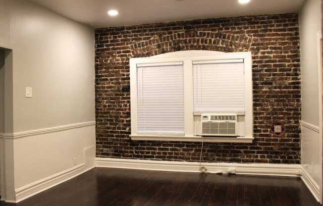 Living room in Brookmore apartments Pasadena CA. Large window with white blinds and trim and air conditioning unit on an exposed brick wall