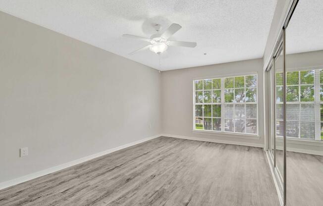 Empty room with light gray walls and a ceiling fan. The floor features light-colored laminate, and there are large windows with blinds allowing natural light. A mirrored closet door is visible on the right side, and the overall space appears clean and minimalist.