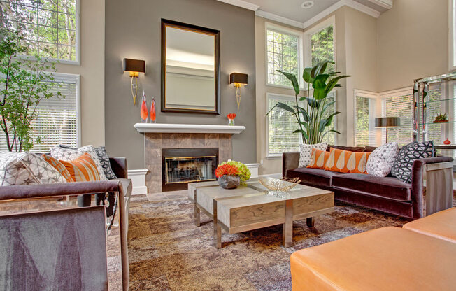 A stylish clubhouse living room here at Aspen Creek showcasing a cozy seating arrangement with plush sofas and patterned accent pillows surrounding a square wood coffee table, centered by a decorative fireplace with a mantel and mirror, tall windows with blinds bringing in natural light, and warm neutral tones accented by greenery and modern décor.