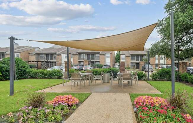 A shaded, outdoor dining area with lush grass and flowers surrounding it at Estelle Creek North Apartments in Irving, TX