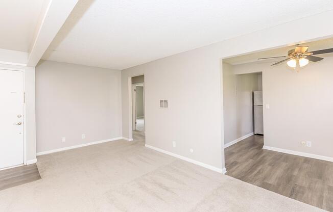 A spacious, well-lit living area featuring neutral-colored walls and carpet. To the left, a door leads to the entrance. There are two openings: one to a room and another to a kitchen area visible in the background. A ceiling fan hangs in the room, adding to the airy feel.