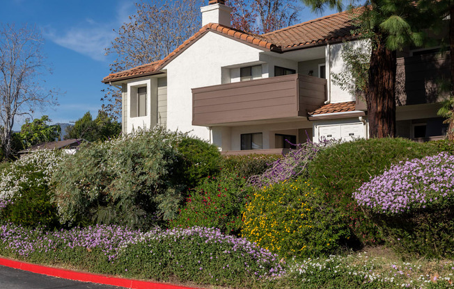 A white house with a red roof and a brown awning is surrounded by a garden with purple flowers.