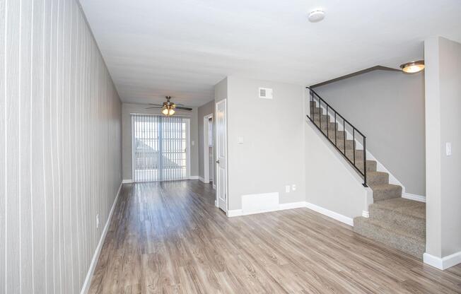 Interior view of a spacious living area featuring light-colored walls, wood-style flooring, and a staircase leading to the upper level. Large windows allow natural light to fill the room, creating an airy atmosphere. A ceiling fan is visible, and there are doors leading to additional rooms.