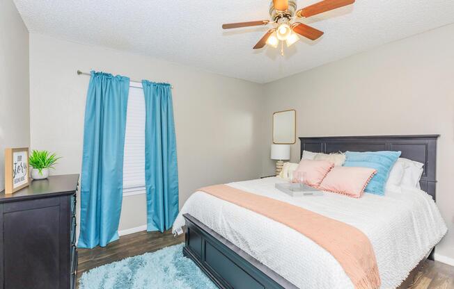 A cozy bedroom featuring a dark wooden bed with a plush white and pastel-colored bedding. There's a modern ceiling fan with light, blue curtains at the window, and a dresser with a decorative plant. A soft blue rug adds warmth to the wooden floor, creating a comfortable and inviting atmosphere.