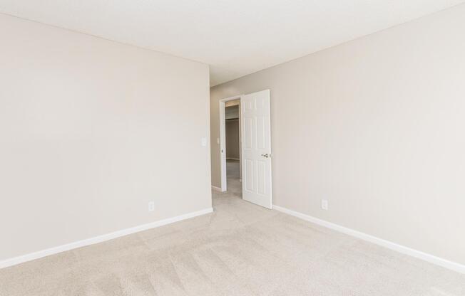 A well-lit, empty room with beige walls and light-colored carpet. A closed door is visible on the left, leading to another space, while an open doorway is seen in the background, suggesting access to additional rooms. The overall atmosphere is neutral and spacious.