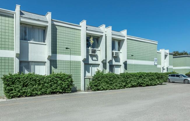 A row of modern green and white buildings with air conditioning units on the windows, set against a clear blue sky. In front of the buildings, there is a well-maintained hedge and a paved road visible. A parked car is seen on the right side of the image.