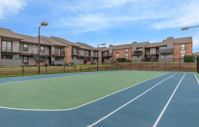 A view of a tennis court surrounded by apartment buildings. The court features a green playing surface with blue lines. In the background, multiple two-story apartments with balconies and landscaped grassy areas are visible under a clear blue sky.