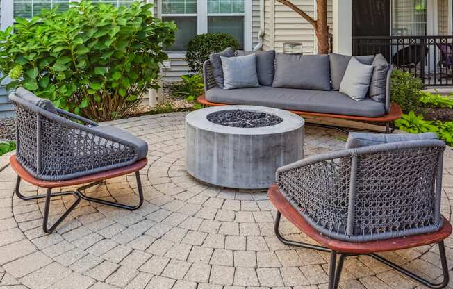 A grey couch with pillows sits on a patio with a fire pit in the middle.