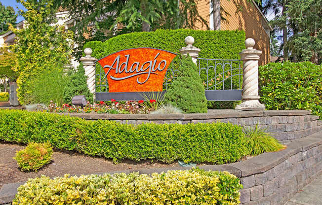 A landscaped community monument sign here at Adagio featuring a decorative stone base, manicured hedges, and colorful flower beds, with a bold Adagio sign framed by ornamental columns and wrought-style fencing, while mature trees and surrounding greenery create a polished, welcoming first impression along the property entrance.
