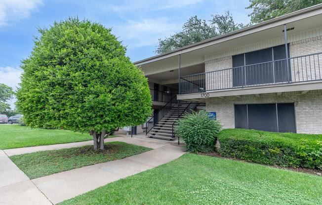 A two-story apartment building with a staircase leading to the second floor. The exterior features brick walls, a well-maintained lawn with a large bushy tree and hedges. The building number is 1700, and the sky is partially cloudy.