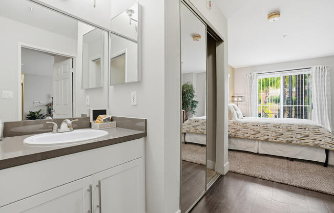 A bathroom with a sink, mirror, and a bedroom with a bed and window.