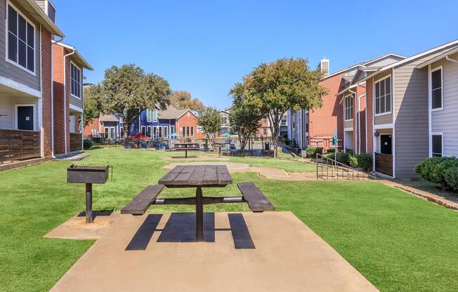 A landscaped courtyard featuring a picnic table on a concrete slab, surrounded by green grass. On either side, there are residential buildings, with some trees and a barbecue grill nearby. The sky is clear, creating a bright and inviting atmosphere.