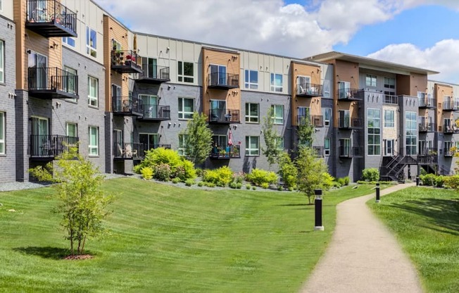 Lakeville, MN Apartments - Lakeside Flats - Building EXterior with Tan and Grey Details, Showcasing Balconies, with Lush Green Lawns, Trees, and Landscaping, and Path
