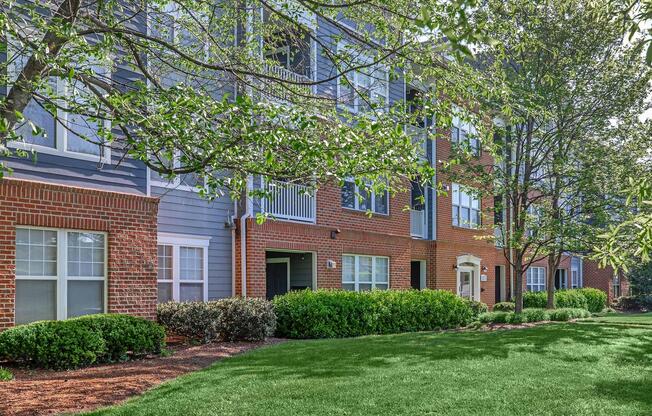 A well-maintained residential building with a mix of brick and blue siding. The facade features multiple windows and balconies, surrounded by lush green grass and neatly trimmed bushes. Several trees provide shade, creating a pleasant outdoor environment.