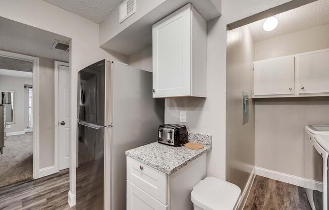 A modern kitchen interior featuring a stainless steel refrigerator, a toaster on a granite countertop, and white cabinetry. The space is well-lit, with a view into an adjoining room and laundry area visible in the background. The flooring is a dark laminate, contrasting with the light-colored walls.
