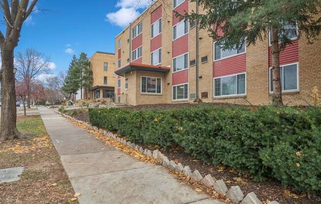 A sidewalk runs alongside a row of bushes and trees in front of a red brick apartment building.