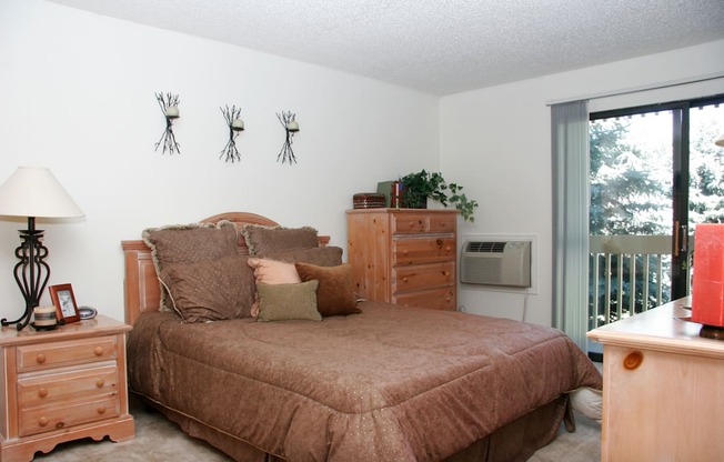 A cozy bedroom here at Eagle Ridge featuring a wood bed frame with layered neutral bedding, matching wood nightstand and dresser, soft carpeted flooring, decorative wall accents above the headboard, and a sliding glass door that brings in natural light and opens to a private balcony overlooking trees.