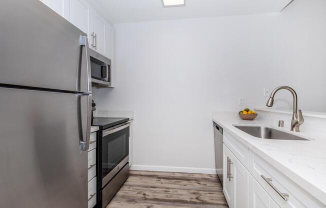 A modern kitchen featuring stainless steel appliances, including a refrigerator, microwave, and oven. The space has white cabinets, a marble countertop, and a large sink. Wood-like flooring adds warmth to the area, while a bowl of fruit sits on the countertop, adding a touch of color.