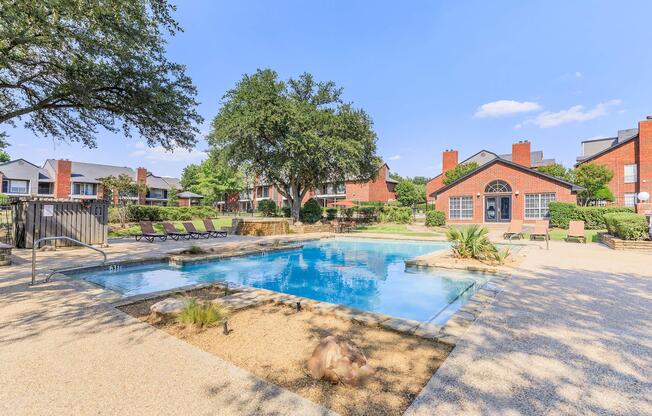 A tranquil pool area surrounded by lush greenery, featuring lounge chairs and a landscaped garden. The pool is clear and inviting, with a stone border. Nearby, two red-brick buildings are visible, and the sky is bright blue with a few clouds, contributing to a relaxing outdoor atmosphere.
