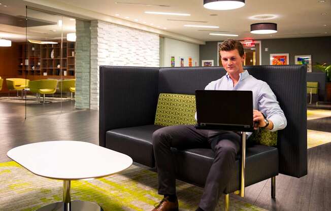 A man in a white shirt and black pants is sitting on a black couch with a laptop on his lap.