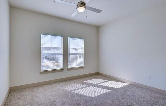 A room with a carpeted floor, two windows with blinds, and a ceiling fan.