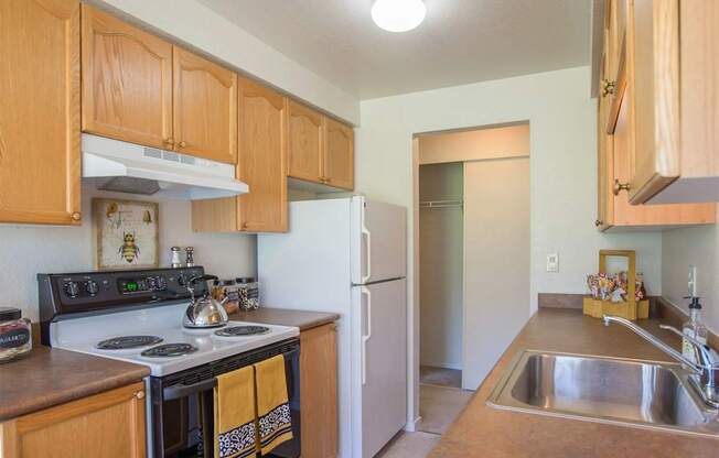 A well-lit kitchen here at Andorra featuring warm wood cabinetry, smooth countertops, and a full appliance setup with a range, refrigerator, and sink, complemented by upper storage, neutral flooring, and a convenient pantry area that adds functionality while maintaining a clean, inviting layout for everyday cooking.