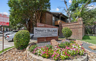 A sign for Trinity Village Apartment Homes is surrounded by flowers and greenery.
