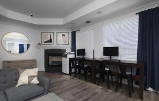 A modern living room featuring a gray sectional sofa with a decorative pillow, a round mirror on the wall, and two framed art pieces. There are two desktop computers on a wooden table next to a printer, with a cozy fireplace on one side and large windows with white blinds covered by blue curtains.