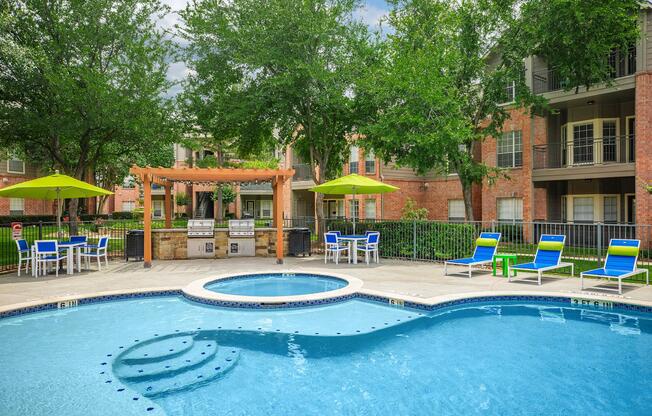 A sunny outdoor pool area featuring a clear blue swimming pool with lounge chairs and green umbrellas. There's a shaded grilling station with a wooden pergola and seating nearby. Surrounding the pool are well-maintained trees and brick apartment buildings in the background.