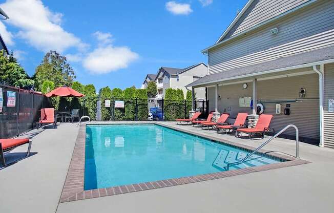 A sunlit outdoor swimming pool here at Allegro featuring a rectangular pool with clear blue water, stainless handrails, and wide concrete decking lined with red cushioned lounge chairs, complemented by a shaded seating area with umbrellas, privacy fencing, landscaped greenery, and surrounding residential buildings under a bright blue sky.