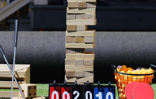 A game of Jenga is set up on a lawn with a scoreboard and ping pong paddles and balls nearby.
