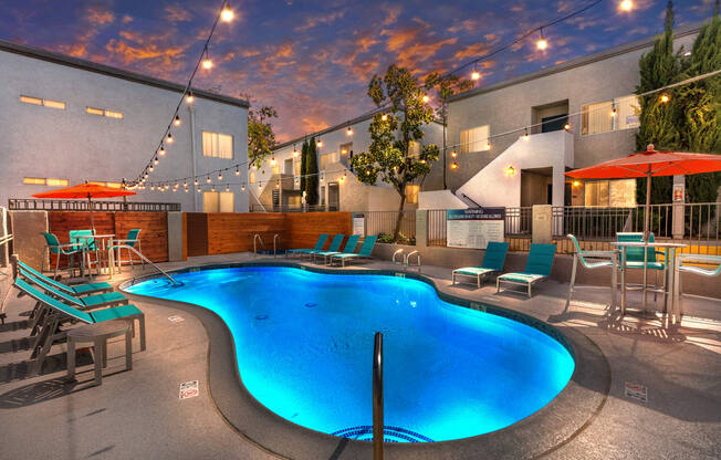 Colton, CA Apartments - Ardella At Reche Canyon - Dusk View Of Swimming Pool With Lounge Seats, Tables, Chairs, Umbrellas, And Cafe Lights.