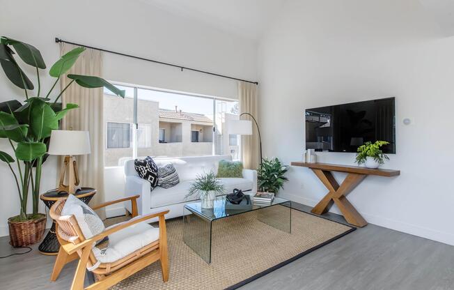 A modern living room featuring a white sofa, a glass coffee table, and a stylish wooden table. There's a large window letting in natural light, a plant in a woven basket, and a TV mounted on the wall. Simple decor creates a minimalist yet inviting atmosphere.