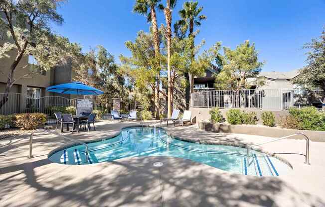 A pool surrounded by trees and chairs under a blue umbrella.