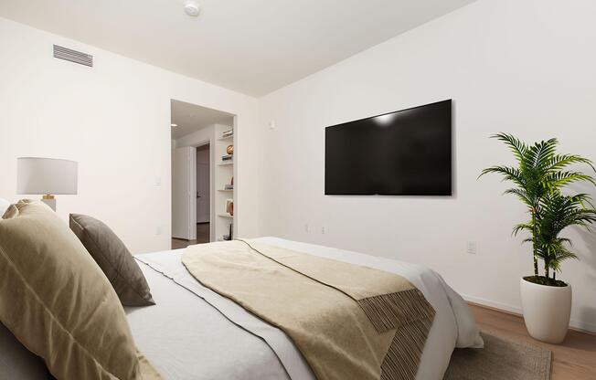 A modern, well-lit bedroom featuring a neatly made bed with beige and white linens, a lamp on a bedside table, a large wall-mounted television, and a potted plant in the corner. A doorway leads to another room, while light-colored walls and wooden flooring create a cozy atmosphere.