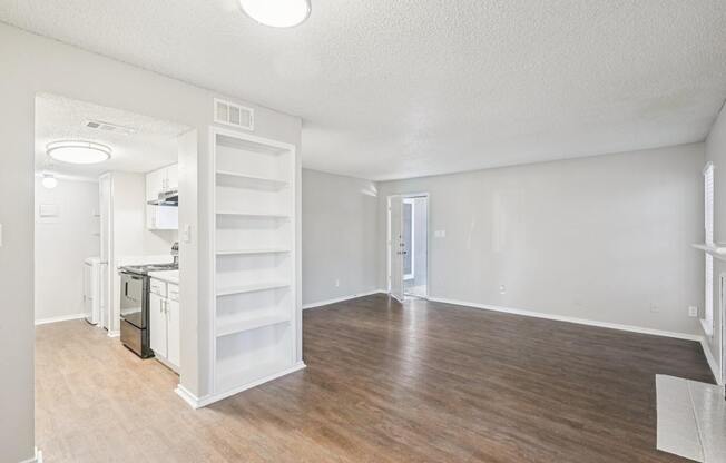 A white room with wood flooring and a kitchen area in the background.