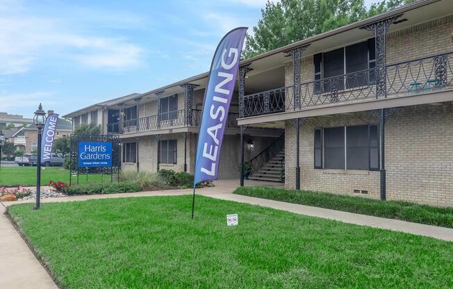 A well-maintained apartment complex with a green lawn. A "Welcome" sign and a large "Leasing" banner are prominently displayed. The building features two stories with outdoor staircases, and there are colorful flower beds at the entrance.
